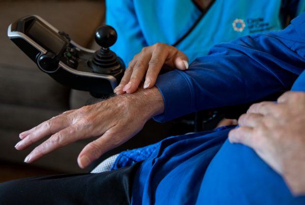 close up of a person dressed in Circle of Care scrubs touching the hand of a person using an electric wheelchair