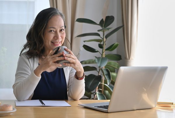 woman at home in front of a laptop, holding a cup and looking at the camera