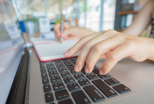 close up of white person's hands on a laptop keyboard and writing in a notebook